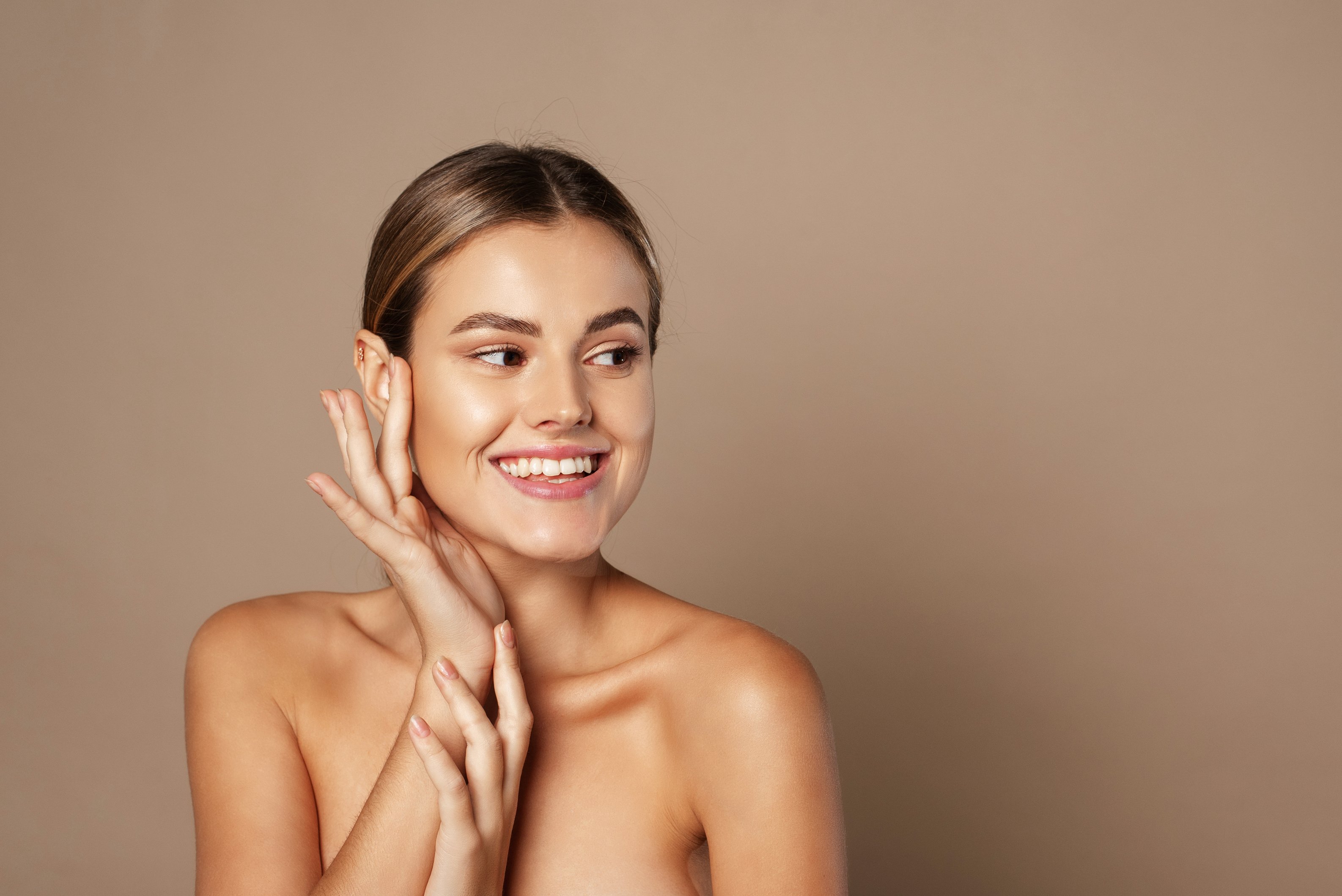 Skin Care. Woman with Beauty Face Touching Healthy Facial Skin. Beautiful Smiling Caucasian Female Model with Natural Makeup Touching Glowing Hydrated Skin on Beige Background